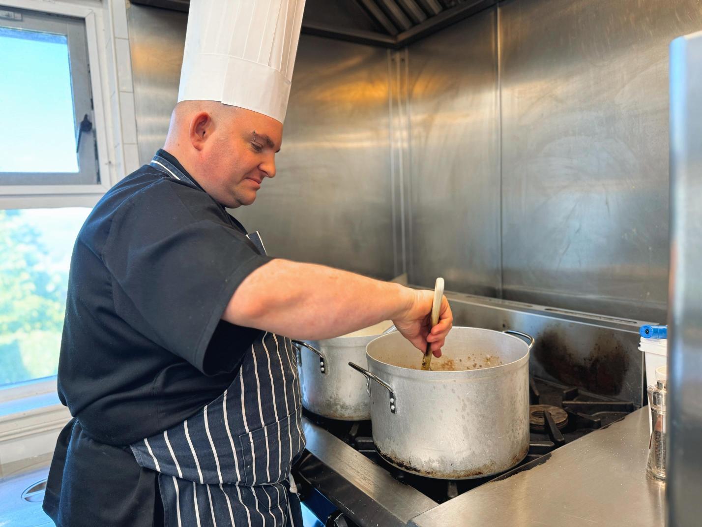 Man wearing a chefs hat an blue and white striped apron, stirring a pot of food in a kitchen.