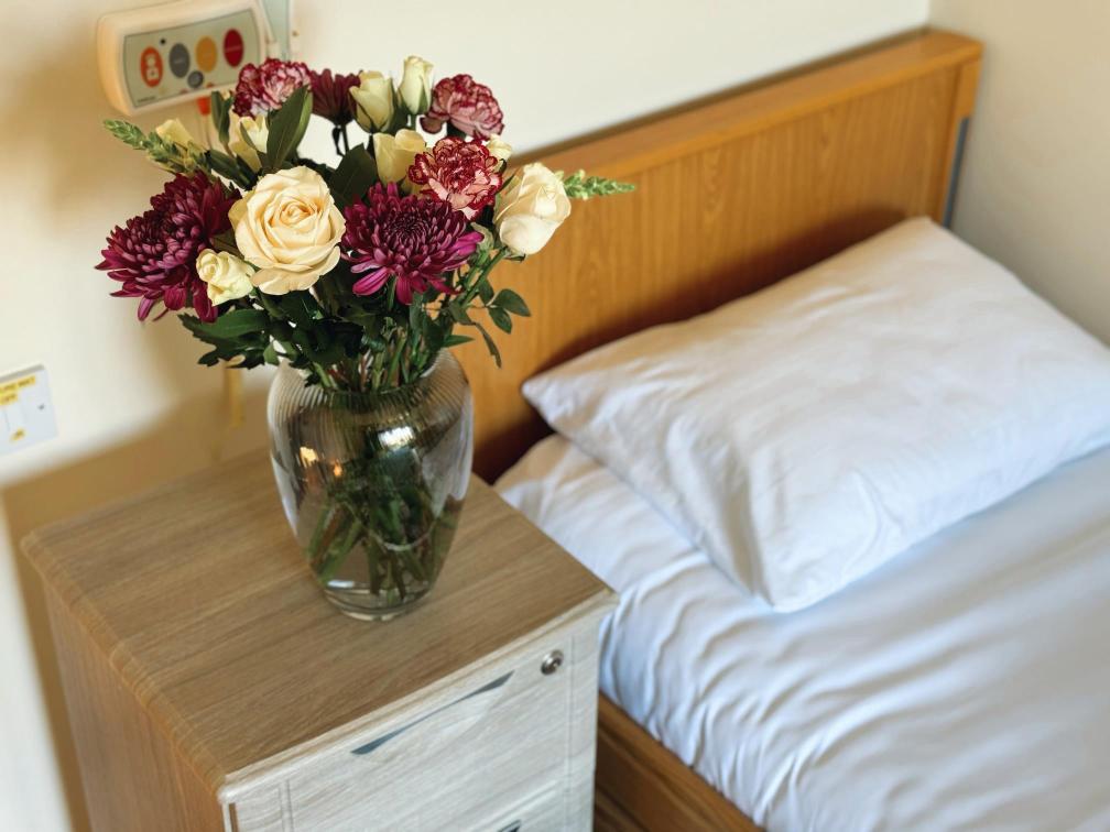 Selection of beautiful flowers in a vase next to a bed.